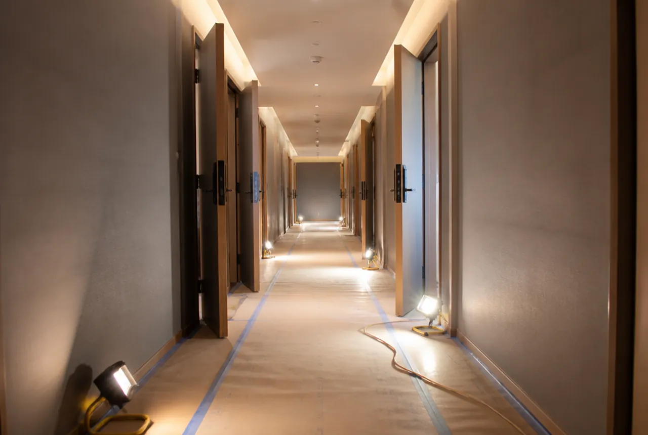 A corridor with open room doors, floor protection and temporary lights along the walls